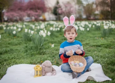 Petite garçon assis portant dans l'herbe porte un panier rempli d'eoufs en chocolat sur ses genoux et un serre-tête oreilles de lapins